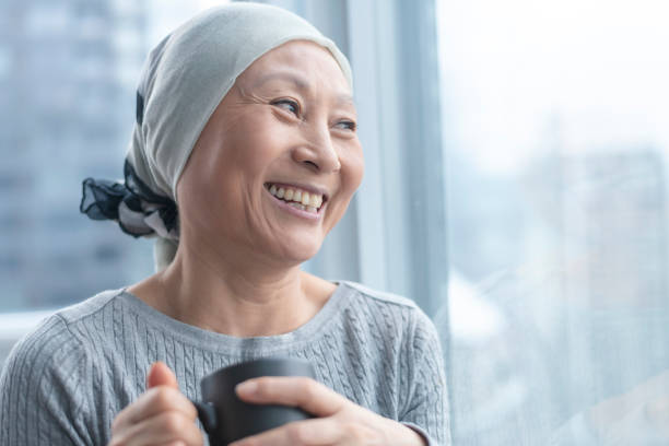 Beautiful Korean woman with cancer looks out window A Korean senior woman with cancer is wearing a scarf on her head. She is standing and holding a cup of tea. The woman leans against a window and smiles with gratitude and hope. chronic illness photos stock pictures, royalty-free photos & images