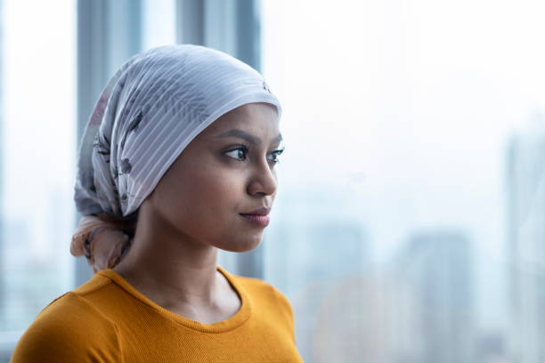 Portrait of beautiful young woman with cancer A Malaysian woman with cancer wears a bandana. She is standing and looking out a window at the city below. She appears contemplative and hopeful. brest cancer hope stock pictures, royalty-free photos & images