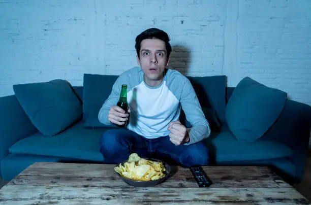 Young man drinking beer at home watching soccer football match or sports game in television at night. Screaming and celebrating goal or victory. In celebration, sports and fans concept.