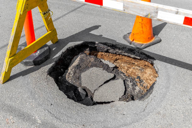 Big Deep Pothole A large, deep pothole in a road. Part of a traffic cone and sawhorse visible blocking the hole. sinkhole stock pictures, royalty-free photos & images