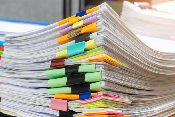 istock Pile of unfinished student's homework assignment on teacher's desk which has a large number of paperwork stacked in archive separated by paper clip with colorful paper. Education and business concept. 1141828161