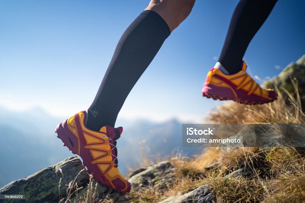 cross country running detail femal legas with compression socks and sport shoes close up legs feet of female cross country trail runner wearing sport compression socks woman running over dry grass high up in mountains side view, focus on shoe Shoe Stock Photo