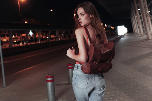 Attractive young girl stands on the pavement with bag on her back and looking to the road side.