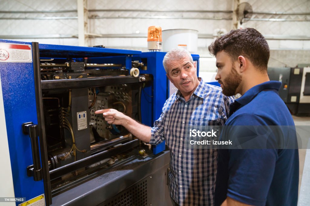 Engineer discussing with trainee over machinery Mature engineer explaining apprentice over injector machine. Instructor is teaching male trainee in industry. They are in factory. 20-24 Years Stock Photo