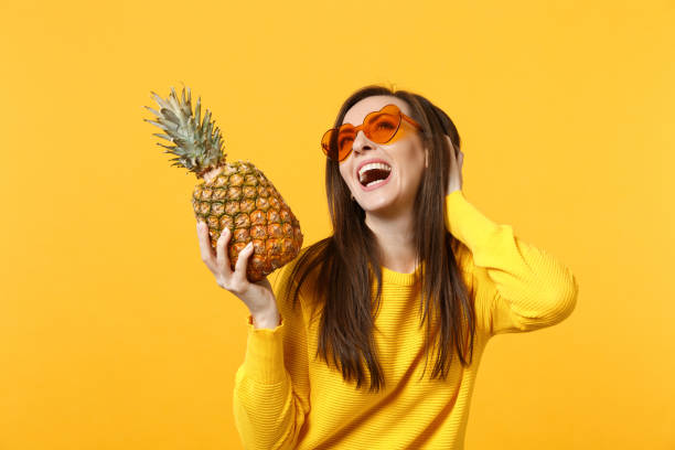 Laughing young woman in heart glasses looking up, holding fresh ripe pineapple fruit isolated on yellow orange background in studio. People vivid lifestyle, relax vacation concept. Mock up copy space mujer joven de risa en gafas de corazón mirando hacia arriba, sosteniendo fruta fresca de piña madura aislada sobre fondo amarillo anaranjado en estudio. personas estilo de vida vivo, relax concepto de vacaciones. mock up espacio de copia.