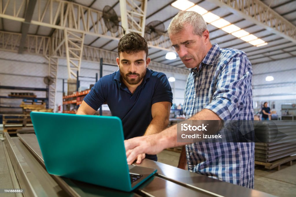 Instructor explaining male trainee over laptop Mature instructor explaining male apprentice over laptop. Trainee is listening to engineer at factory. They are in industry. Instructor Stock Photo