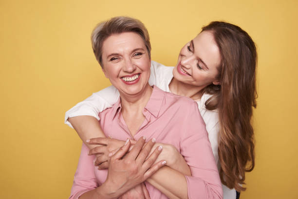 Beautiful Caucasian girl is hugging her mother while posing for camera Waist up of brunette smiling young girl looking at her mummy while standing behind. Old woman is looking at camera while situating against orange background. Trusting relationship concept adult offspring photos stock pictures, royalty-free photos & images