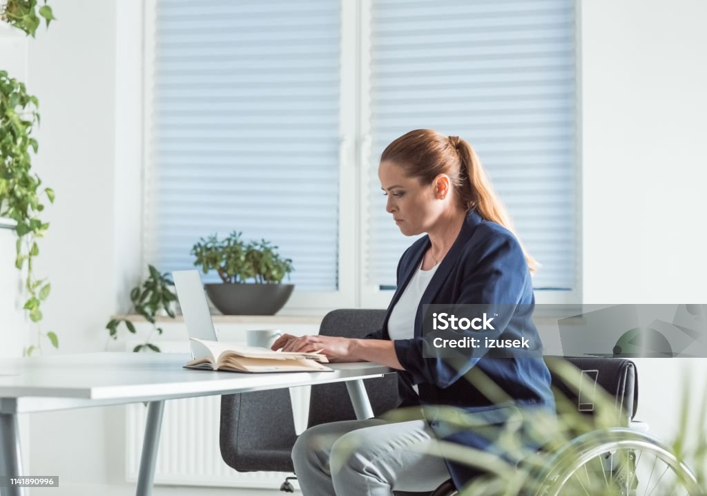 Disable businesswoman reading book at desk Disable businesswoman reading book at desk. Female professional is sitting on wheelchair in office. She is in businesswear. Business Stock Photo