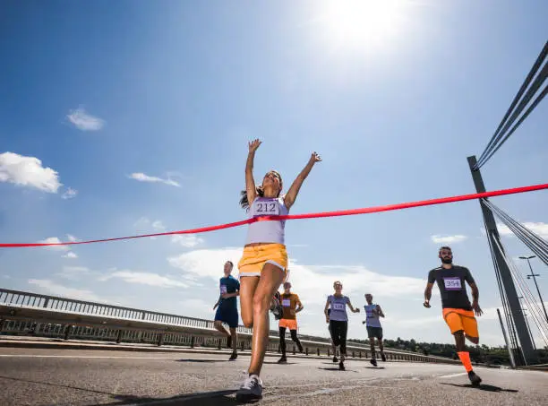 Low angle view of female marathon runner celebrating victory while crossing the finish line with raised arms. Copy space.