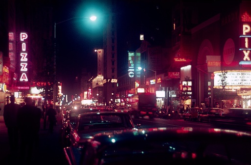 New York City, NY, USA, 1962. The Broadway with its cinemas, shops, theaters, entertainment shows and restaurants by night. Furthermore: neon signs, cars, traffic and passers-by.