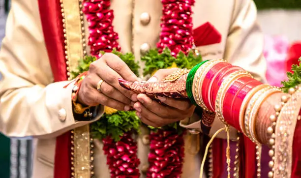 Photo of Engagement Ring ceremony- Indian Hindu male putting ring on bride's decorated finger. Couple is well attired as per traditional Indian Hindu wedding. Groom wearing Jodhpuri suit and floral garland.