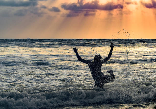 chico adolescente en vacaciones, divirtiéndose y celebrando la vida en una playa. él está saltando libremente para bucear en las olas del mar con sus manos y piernas en el aire. hermosa puesta de sol en el fondo. - florida mid air miami florida people fotografías e imágenes de stock