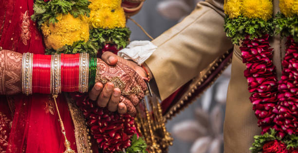 Indian Hindu couple wearing traditional Indian dress and floral garland, holding each other hands during their marriage promising blissful life. Beautiful Henna Mehndi art and bangles on birde hands Beautiful photo of a Newly Married Indian Couple holding hands and promising each other for living together merrily for all their life. bangle photos stock pictures, royalty-free photos & images