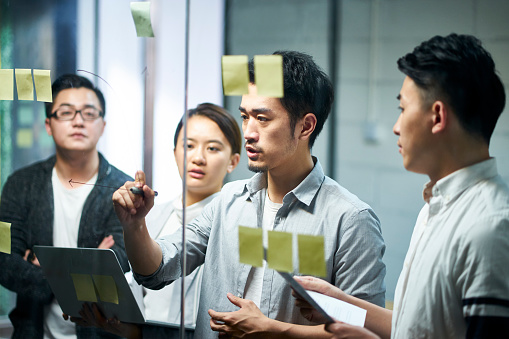 young asian entrepreneur of small company drawing a diagram on glass during team meeting discussing and analyzing business situation in office.