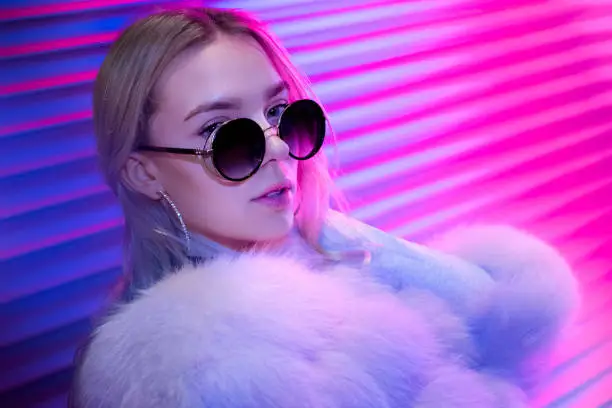 Teen hipster girl in stylish glasses and fur posing on street neon light wall background, female teenager fashion model woman with beautiful face looking at camera in city night light glow, back to 80s, portrait