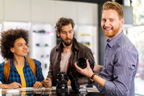 istock Store seller looking at a camera and smiling while helping two young photographers choose various pieces of photographic equipment in a camera shop 1141930953