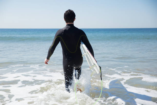 istock Back view of man carrying surfboard and walking into ocean water 1141945385