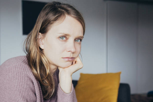Upset young woman sitting on sofa at home, looking at camera Upset young fair-haired Caucasian woman in purple sweater, grey home trousers sitting on grey sofa in living room, looking at camera. Lifestyle concept medium shot stock pictures, royalty-free photos & images