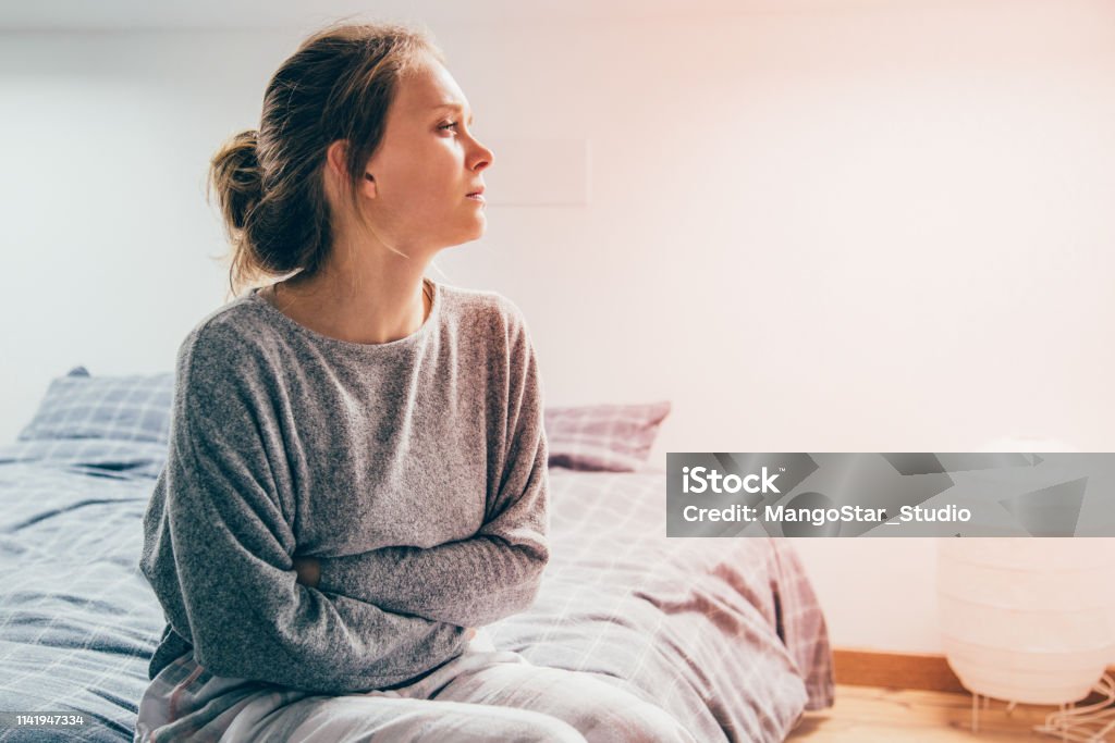 Woman with hands on stomach suffering from pain, looking aside Tired sick woman in grey homewear sitting on bed, keeping hands on stomach, suffering from pain, looking aside. Illness, stomach ache concept Women Stock Photo