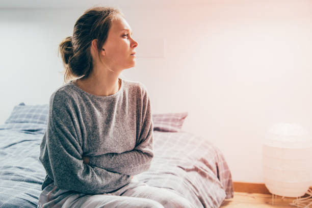 Woman with hands on stomach suffering from pain, looking aside Tired sick woman in grey homewear sitting on bed, keeping hands on stomach, suffering from pain, looking aside. Illness, stomach ache concept medium shot stock pictures, royalty-free photos & images