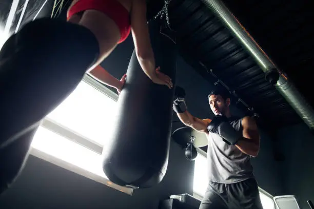A female holding a boxing punch bag as young man punches it with boxing gloves in a gym.
