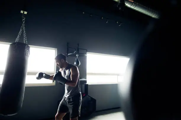 A latin male standing in a gym wearing boxing gloves and punching a boxing glove.