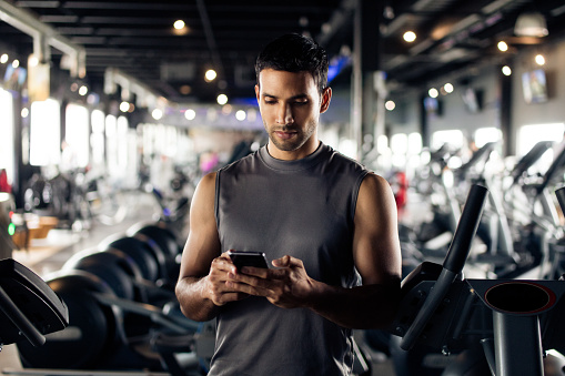 A young latin man standing in a gym next to the treadmills using his cellphone to text.