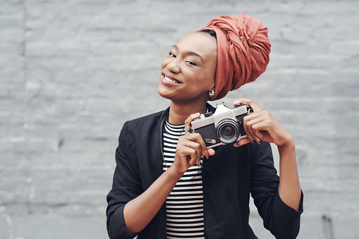 Cropped portrait of an attractive young businesswoman posing with her camera against a grey brick wall outside