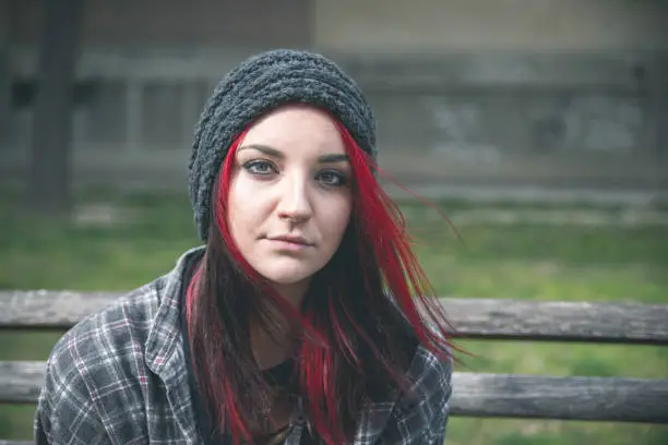 Photo of Young beautiful red hair girl sitting alone outdoors on the wooden bench with hat and shirt feeling anxious and depressed after she became a homeless person close up portrait