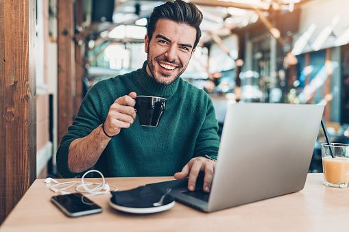 Smiling young man drinking coffee and using laptop in cafe