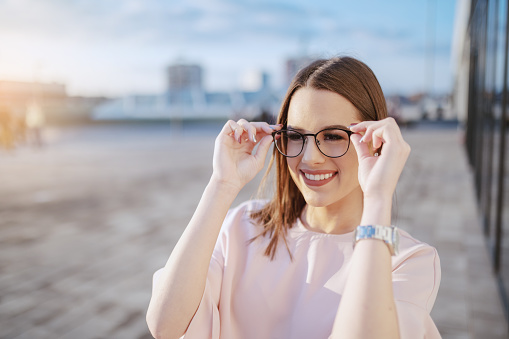 Charming smiling Caucasian brunette dressed elegant putting eyeglasses while standing outdoors.