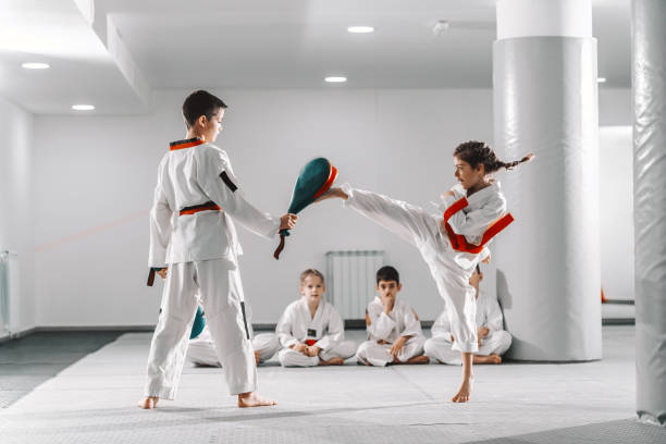 caucasain boy and girl in doboks having taekwondo training at gym. girl kicking while boy holding kick target. in background their friend sitting with legs crossed and watching them. - weight class imagens e fotografias de stock