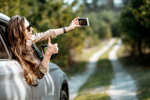 Young woman enjoying the trip, photographing out the window with phone on a picturesque road in the woods