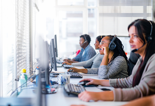 Customer service representatives using computers at desk. Focus is on African woman talking on headset. They are working in call center.