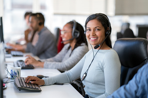 Portrait of smiling businesswoman wearing headset while using computer. Confident female operator is working with colleagues at desk. They are in call center.