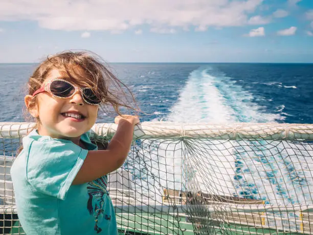 little girl with sunglasses caught at the railing of the cruise ship in which she travels smiles at the camera, the wind moves her hair and the white foam from the ship's wake moves through the ocean