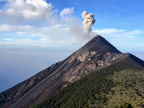 A close up view of mount fuego volcano during the day outside of Antigua, Guatemala.  Smoke and ash are rising out of the top or crater of the volcano.  The side is black from previous eruptions.