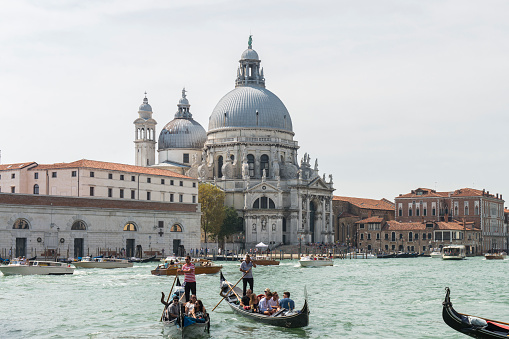 Gondolas with tourists sailing in the Grand Canal near Santa Maria della Salute (Saint Mary of Health) church at Venice, Italy. The Grand Canal is an ancient waterway that goes from the Saint Mark Basin to a lagoon near the Santa Lucia rail station and is lined with buildings constructed from the 13th to the 18th centuries by wealthy Venetian families.\nThe city of Venice is an Unesco World Heritage site and it is visited daily by thousands of tourists from all over the world. The gondola tour is a preferred attraction for the tourist at Venice.