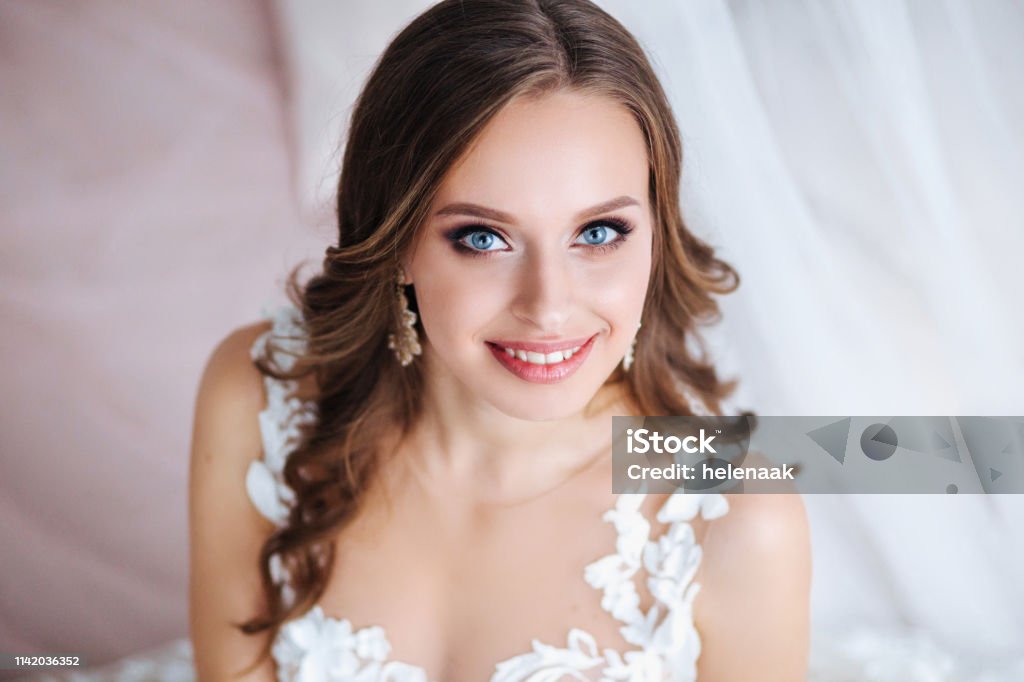 Beautiful bride with long curly hair in elegant wedding dress and diadem smiling in room in the wedding morning Beautiful bride with long hair in elegant wedding dress and diadem smiling in room in the wedding morning Bride Stock Photo