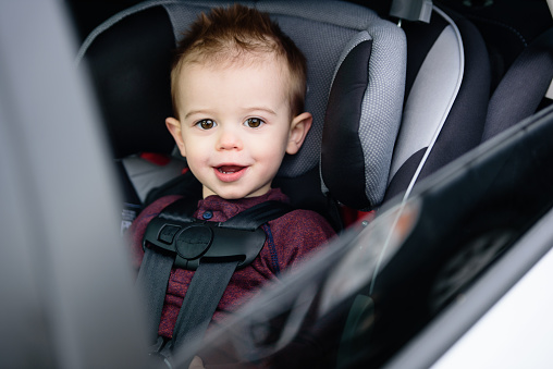 Smiling toddler boy strapped into car seat in car. Looking through the window at the camera.