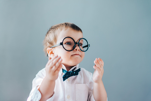 Cute little boy wearing smart outfit with a bow tie and eyeglasses, clapping his hands. Plain grey background.