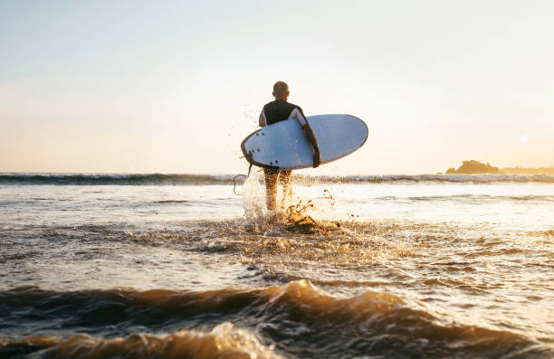 istock Young man Surfer taking surfboard and comming with long surf board to waves on the evening sunset sky background. Active holidays spending concept. 1142087999