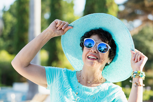 A senior Hispanic woman in her 60s wearing sunglasses and a wide-brimmed hat on a sunny day. She is on vacation at a tropical resort, standing on a patio, smiling and looking up at the sky. A building, palm trees and clear blue skies are visible in the reflection in the sunglasses.
