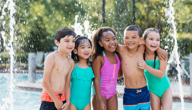 Multi-ethnic children at water park A multi-ethnic group of children 6 to 9 years old having fun at a water park. They are wearing swimsuits, standing by a swimming pool with water falling and splashing behind them. male swimsuit standing arm around stock pictures, royalty-free photos & images