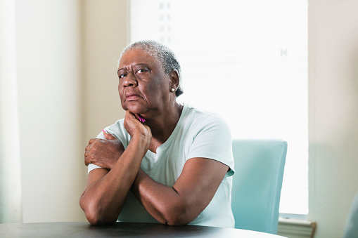 A lonely senior African-American woman in her 70s sitting at home at a table by a window. She is looking at the camera with a serious expression, elbows on the table and head leaning on her hand.