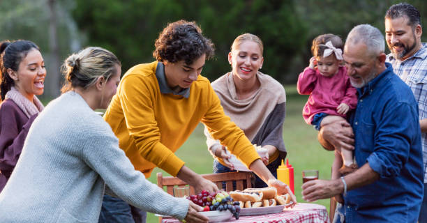 Multi-generation Hispanic family having backyard cookout A multi-generation Hispanic family having a backyard cookout. The large family ranges in age from 16 months to 50s, including grandparents, parent, grandchild, sons, and daughters. They are putting food down on a table, smiling and conversing. A mature man in his 50s is holding his granddaughter, a 16 month old baby. The teenage boy in the orange sweater is the girl's uncle. big family table stock pictures, royalty-free photos & images