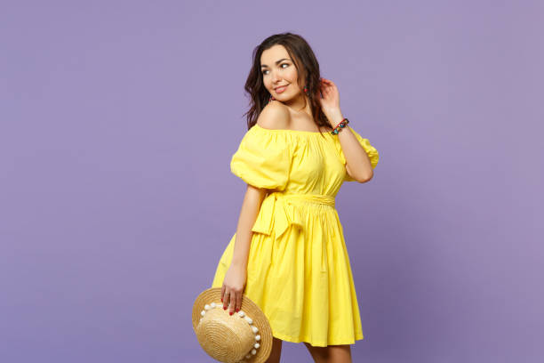 Portrait of pretty young woman in yellow dress holding summer hat looking aside isolated on pastel violet wall background in studio. People sincere emotions, lifestyle concept. Mock up copy space retrato de mujer joven bonita en vestido amarillo sosteniendo sombrero de verano mirando a un lado aislado en el fondo de la pared violeta pastel en estudio. personas emociones sinceras, concepto de estilo de vida. mock up espacio de copia.