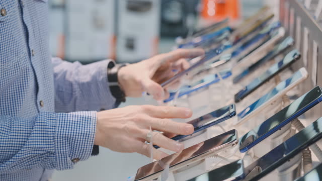 Close-up of a man's hand rotates and inspects the design of a new smartphone in a modern electronics store. Take the phone before buying