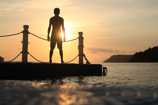 Asian man standing and jumping on floating pier at sunrise , Silhouette body of asian people early morning on the beach by the sea.