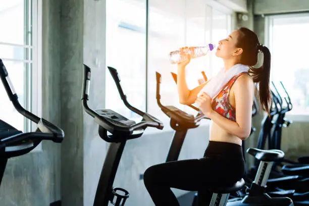 Gym woman working out drinking water smiling happy.young asian girl exercising on bicycle in fitness center.sport and Healthy concept. Workout with happiness ideas.